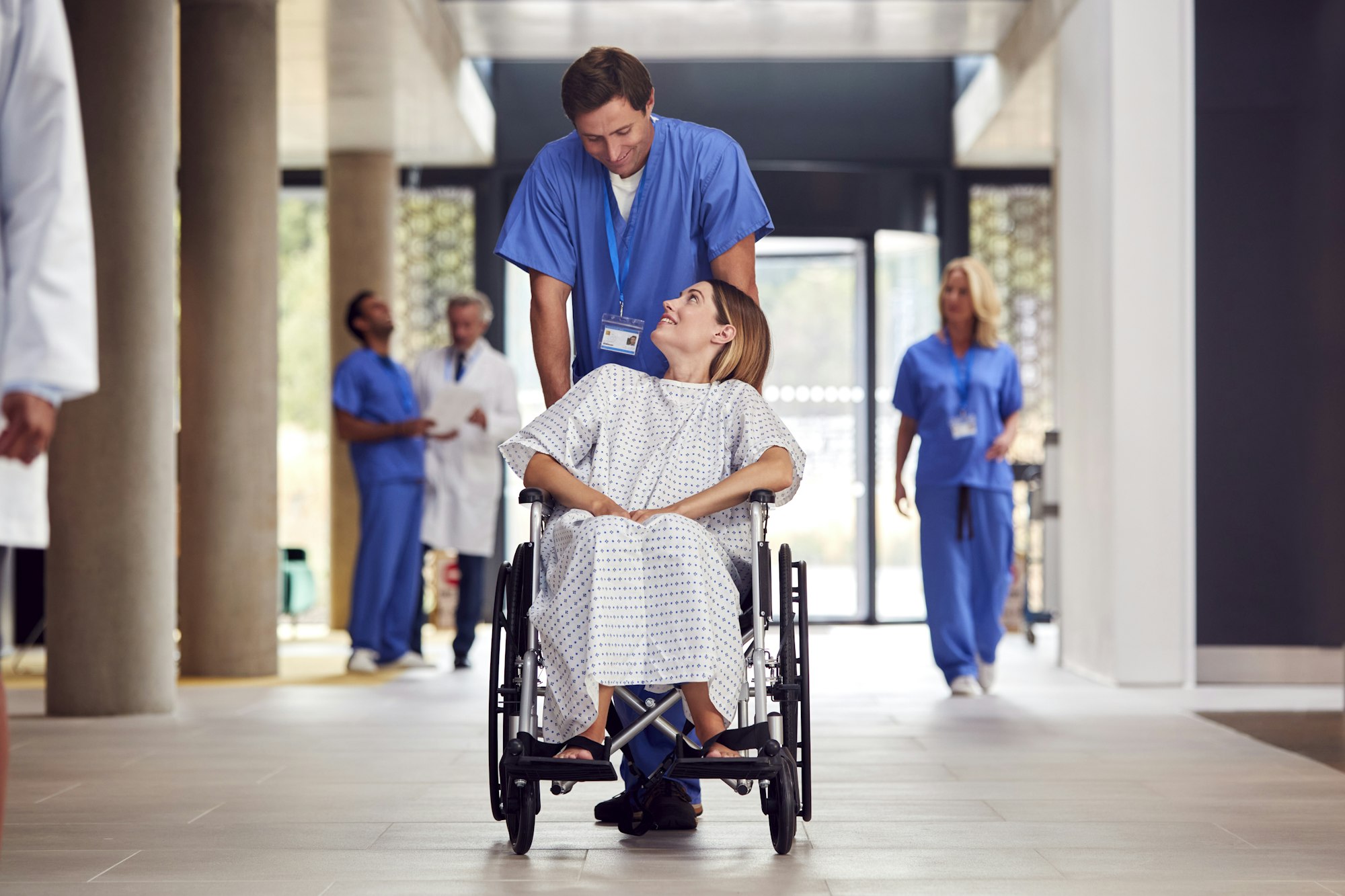 Male Nurse Wearing Scrubs Pushing Female Patient In Wheelchair Through Hospital Building